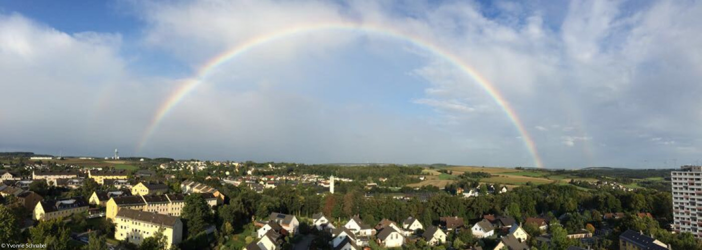 Im Hintergrund blauer Himmel und weiße Wolken, in der Mitte die Lutherkirche, umgeben von Häusern, darüber leuchtend der Regenbogen