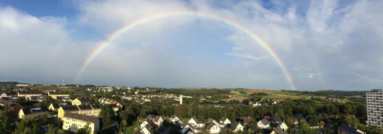 Im Hintergrund blauer Himmel und weiße Wolken, in der Mitte die Lutherkirche, umgeben von Häusern, darüber leuchtend der Regenbogen